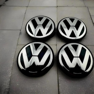 May include: Four black and silver hubcaps with the Volkswagen logo. The hubcaps are circular with a black background and a silver "VW" emblem in the center. The hubcaps are arranged in a square formation.