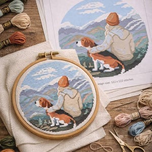 May include: A cross-stitch design in a wooden hoop, depicting a person and a dog gazing at mountains, with the text "Serenity in the mountains." The image includes embroidery threads, scissors, and a printed cross-stitch chart.