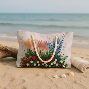 May include: A white tote bag with a floral design featuring red roses, blue and white flowers, and green foliage. The bag has rope handles and gold-coloured eyelets. The background shows a beach scene with sand, seashells, and a rolled-up towel.
