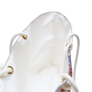 May include: White tote bag with rope handles and gold-coloured eyelets. The interior is white, and the exterior features a floral pattern with red, blue, and green accents. The bag is designed for carrying items.