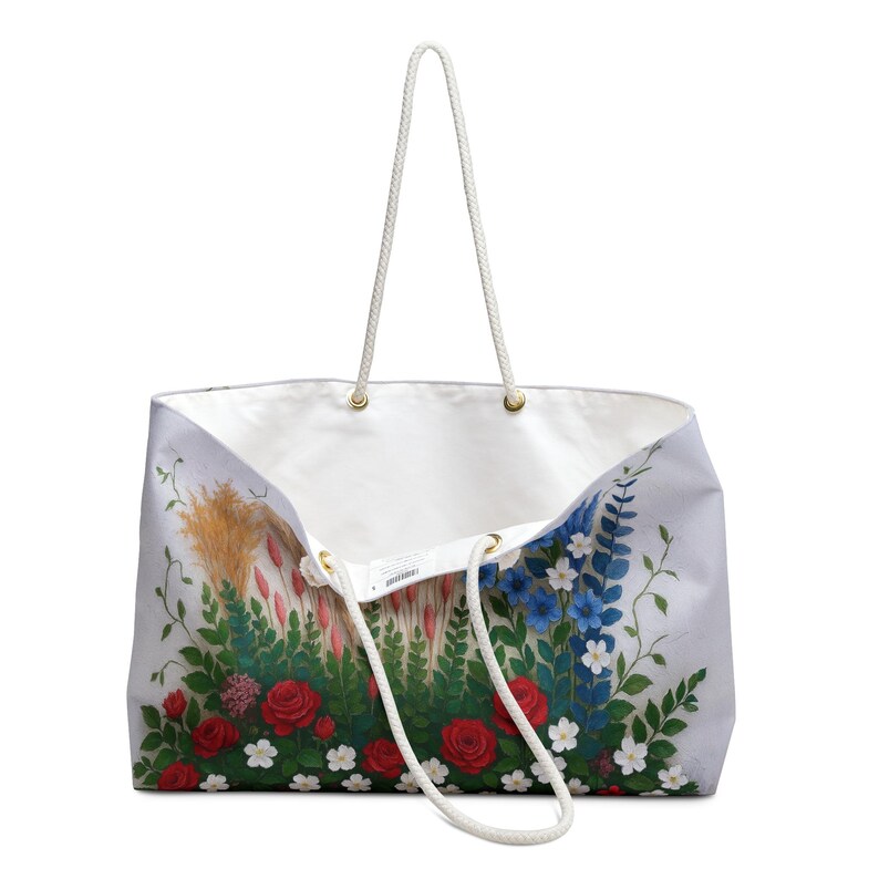 May include: White tote bag with a floral design featuring red roses, blue and white flowers, and green foliage. The bag has white rope handles and gold-coloured eyelets. The interior is white.