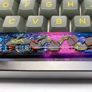 Könnte beinhalten: Eine benutzerdefinierte Tastatur-Leertaste mit einem kosmischen Design und einer schlangenartigen Kreatur. Die Taste hat ein dunkelblaues, lila und rosa Galaxie-Design mit einer grauen, gelben und roten Kreatur.