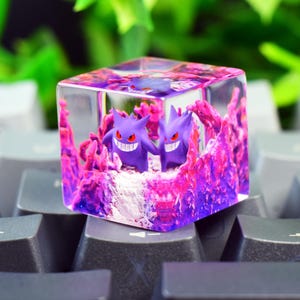 Könnte beinhalten: Eine transparente Tastenkappe aus Kunstharz in Würfelform mit zwei violetten Gengar-Figuren im Inneren, umgeben von strukturierten rosa und violetten felsartigen Formationen. Der Würfel liegt auf einer dunkelgrauen Computertastatur mit weißen Symbolen.
