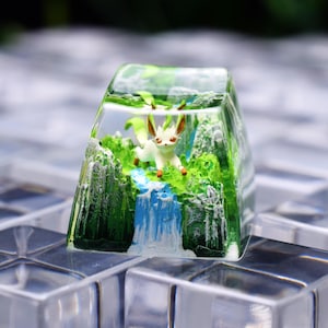 May include: A clear resin mechanical keyboard keycap featuring a miniature diorama of a white and tan creature with large ears standing in a green forest landscape with a blue waterfall and rocky cliffs encased within the transparent material.