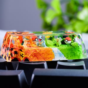 May include: A rectangular resin keyboard keycap featuring a miniature landscape with a lava-like orange and red section on the left and a green grassy section on the right, containing small character figures and two red and white spherical icons.