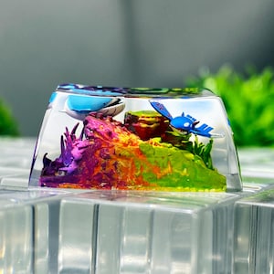 May include: A clear resin artisan keycap featuring a miniature underwater scene with a vibrant, multi-colored rocky formation in shades of purple, orange, and green, accompanied by two small blue aquatic creature figures inside the transparent casing.