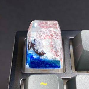 Pode incluir: Uma tecla de teclado personalizada com uma cena oceânica em miniatura. A tecla transparente tem uma base azul oceano, ondas brancas e uma cordilheira vermelha e branca. A tecla é retangular e está em um teclado cinza.