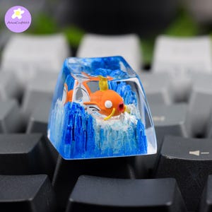 May include: A custom artisan keycap featuring a vibrant orange and white cartoon fish character set in a clear resin block. The keycap has a blue water effect and is set against a gray and black keyboard.