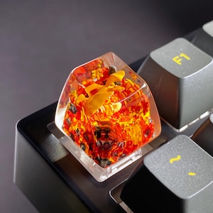 May include: A custom keyboard keycap with a clear resin exterior. The keycap features a fiery, lava-like design in red, orange, and black, with a yellow accent. The keycap is installed on a black keyboard.