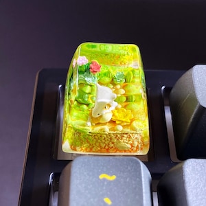 May include: A custom keyboard keycap with a miniature landscape scene. The keycap is transparent with a green and yellow colour scheme, depicting a small creature among flowers and foliage. The keycap is set on a black keyboard.