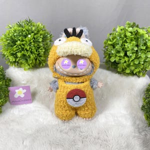 Puede incluir: Muñeco de peluche con un disfraz amarillo y gris con un gráfico rojo y blanco. El muñeco tiene una cara sonriente con ojos morados y una capucha amarilla. El muñeco está sobre una superficie blanca con plantas verdes en el fondo.