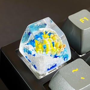 Könnte beinhalten: Eine individuelle Tastaturkappe mit einem gelb-blauen Meereskreaturen-Design, das in klarem Harz eingeschlossen ist. Die Kappe ist von blauen und weißen Wellen umgeben und befindet sich zwischen grauen Tastaturtasten.