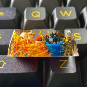 May include: A custom resin keycap featuring a split design with a fiery orange and blue water theme. It includes miniature figurines of a Pokémon character, a Pokéball, and other decorative elements. The keycap is set against a black keyboard.