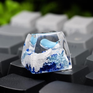 May include: A clear resin mechanical keyboard keycap featuring a miniature light blue whale swimming above textured white and deep blue waves. The keycap is shaped like a standard square key and is positioned among other dark gray keyboard keys.