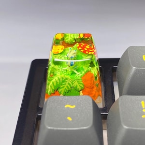 Puede incluir: Una tecla de teclado personalizada con un diseño detallado y colorido. La tecla muestra una pequeña criatura verde en medio de un follaje verde vibrante, acentos naranjas y setas rojas con lunares. La tecla está sobre un teclado gris.