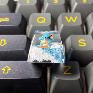 Peut inclure: Un bouton de clavier personnalisé représentant une tortue bleue portant des lunettes de soleil et surfant sur une vague. Le bouton est transparent avec un motif de vague bleu et blanc, sur un clavier gris.