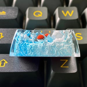 May include: A custom artisan keycap depicting a blue and white scene with cartoon characters and a red and white ball. The keycap is set against a black keyboard.