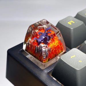 May include: A custom artisan keycap with a purple dragon figurine set in a fiery, lava-like scene. The keycap is encased in clear resin and is mounted on a grey keyboard.