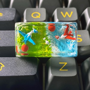 May include: A custom keycap featuring a split design with two distinct scenes. One side showcases a blue and white flying creature with a red sphere against a green background. The other side displays a red and white flying creature with a red sphere against a blue background.