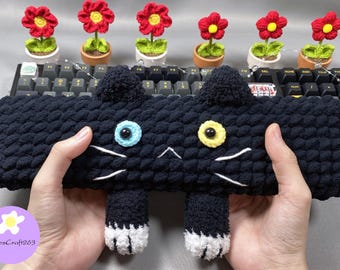 May include: A handmade black cat-shaped crochet item with blue and yellow eyes, white whiskers, and white-tipped paws. The cat is held in front of a keyboard and several small potted flowers with red blooms.
