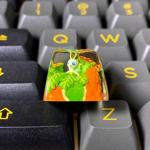 May include: A custom keycap featuring a small, white cartoon character with blue eyes, set within a clear resin block. The keycap is surrounded by green foliage and orange terrain, resting on a gray keyboard.
