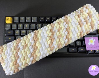 May include: A rectangular crochet wrist rest featuring a textured, bobble-stitch pattern in alternating diagonal stripes of white, pale yellow, and light tan yarn, positioned atop a black mechanical keyboard with a small purple square tag reading AriesCraft263.
