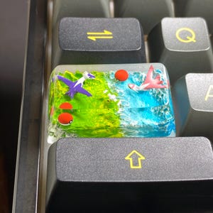 May include: A custom keyboard keycap with a miniature landscape. It features a purple and white flying creature, a pink flying creature, and red and white spheres. The keycap has a green and blue background, surrounded by black keyboard keys.