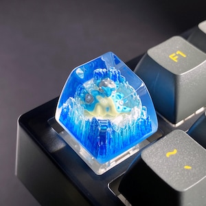 Puede incluir: Una tecla de teclado personalizada con un diseño azul y blanco de una criatura marina. La tecla está hecha de resina transparente, con un diseño detallado y en capas. La tecla está en un teclado negro.