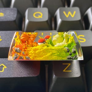 May include: A custom artisan keycap featuring a vibrant scene with orange and yellow flames, a fiery fox-like creature, and a green landscape with a white and yellow creature. Two red and white Poké Balls are also present.
