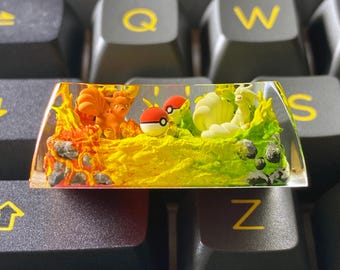 May include: A custom artisan keycap featuring a vibrant scene with orange and yellow flames, a fiery fox-like creature, and a green landscape with a white and yellow creature. Two red and white Poké Balls are also present.