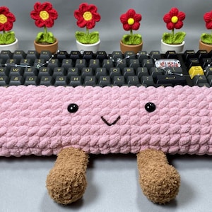 Peut inclure: Un repose-poignet de clavier rose et moelleux avec un visage souriant et deux pieds marron et flous. Au-dessus du clavier, six petites plantes en pot avec des fleurs crochetées rouges et jaunes. Le clavier a des touches noires.