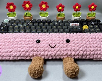 May include: A pink, plush keyboard wrist rest with a smiling face and two brown, fuzzy feet. Above the keyboard are six small potted plants with red and yellow crocheted flowers. The keyboard has black keys.