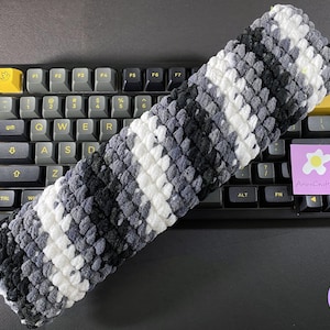 Peut inclure: Un repose-poignet pour clavier à rayures grises, blanches et noires, posé sur un clavier mécanique. Le repose-poignet est fait d'une matière douce et texturée. Un autocollant avec le texte "AriesCraft263" est visible.