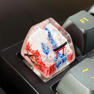 May include: A custom keyboard keycap featuring a miniature Japanese landscape. The keycap is clear with a mountain scene, a red torii gate, and red and white cherry blossom trees. The keycap is on a black keyboard.