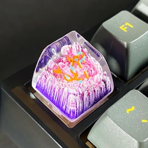 May include: A custom keyboard keycap with a mountain landscape design. The keycap features a clear resin exterior with a pink and purple mountain range and an orange and blue figure. The keycap is on a black keyboard.