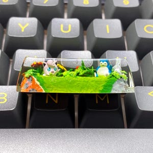 Peut inclure: Un keycap personnalisé avec un diorama miniature. Il présente deux personnages de dessins animés, un rose et un bleu, dans un paysage vert avec un champignon et du feuillage. Le keycap transparent est posé sur un clavier noir.