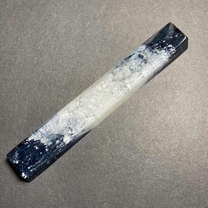 Puede incluir: Un bloque rectangular de resina para bolígrafo con un diseño jaspeado de remolinos azul marino profundo y blanco translúcido. La superficie tiene un acabado brillante con patrones complejos de pigmento blanco en la resina azul.