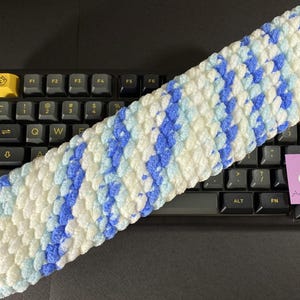 May include: A handmade, rectangular crochet headband in a textured stitch pattern. The headband features a repeating pattern of white, light blue, and blue stripes. The headband is resting on a black keyboard.