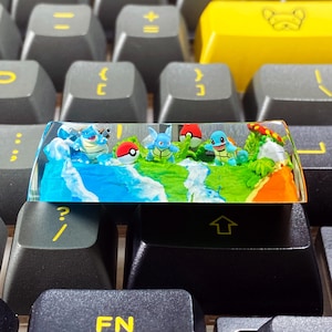 May include: A custom artisan keycap featuring a miniature diorama of a water-themed landscape with Pokemon characters and Poke Balls. The keycap is clear with a colorful design, set against a gray keyboard.