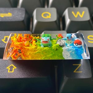 Op de afbeelding: Een aangepaste keycap met een kleurrijk diorama met miniaturen van Charmander, Bulbasaur, Squirtle en een Poké Ball. De keycap is geplaatst op een toetsenbord, met de letters Q, W en Z zichtbaar.