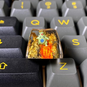 May include: A custom keyboard keycap with a green and blue cartoon character on a fiery, gold-coloured base. The keycap is transparent, encased in resin, and sits on a grey keyboard.