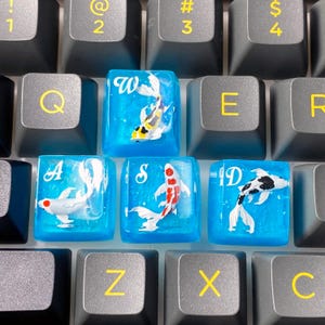 May include: Close-up of custom keyboard keycaps. The keycaps are blue with koi fish designs. The letters W, A, S, and D are visible on the keycaps. The surrounding keys are gray with yellow lettering.