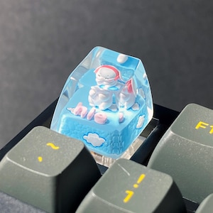 Puede incluir: Una tecla de teclado personalizada con un exterior de resina transparente. En el interior, una base azul presenta nubes blancas y la palabra "hi" en rosa. También se ven dos figuras blancas con detalles en rosa.