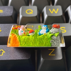 May include: A custom keycap featuring a miniature landscape with cartoon characters. The keycap has a clear resin base with a green grassy area, an orange cliff, and a gray rock formation. The characters include a pink Jigglypuff, a red and white Poké Ball, and a blue Snorlax.