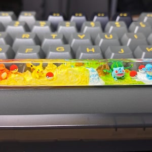 May include: A custom artisan keycap featuring a colorful landscape with various Pokemon characters. The keycap is a rectangular shape and is set on a gray keyboard. The design includes fire, yellow, green, and blue sections.