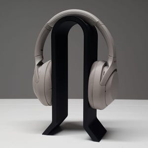 May include: Light grey over-ear headphones displayed on a black headphone stand. The stand has a curved top and a rectangular base. The headphones are a light grey colour.