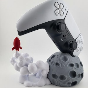 May include: A white and black games controller sits on a grey moon-shaped base with a 3D-printed red rocket taking off. The base has a textured surface with craters. The controller is a modern design.