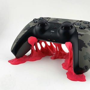 May include: A black and camouflage-patterned video game controller on a bright red, stylized stand. The stand appears to be a 3D-printed design, adding a unique aesthetic to the gaming accessory. The controller has black buttons and joysticks.