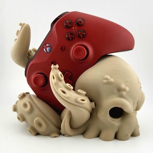 May include: A red games controller sits on a beige octopus-shaped stand. The controller has black buttons and a black directional pad. The octopus stand has multiple tentacles and a detailed head. The logo "3DCRAFT CODE" is visible in the bottom left corner.