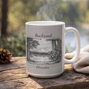 May include: A white ceramic mug featuring a black and white sketch of a mountain landscape, a lake, and a hanging flower basket. The text "Backyard" is printed above the illustration, and the word "Dreams" is printed below the illustration.